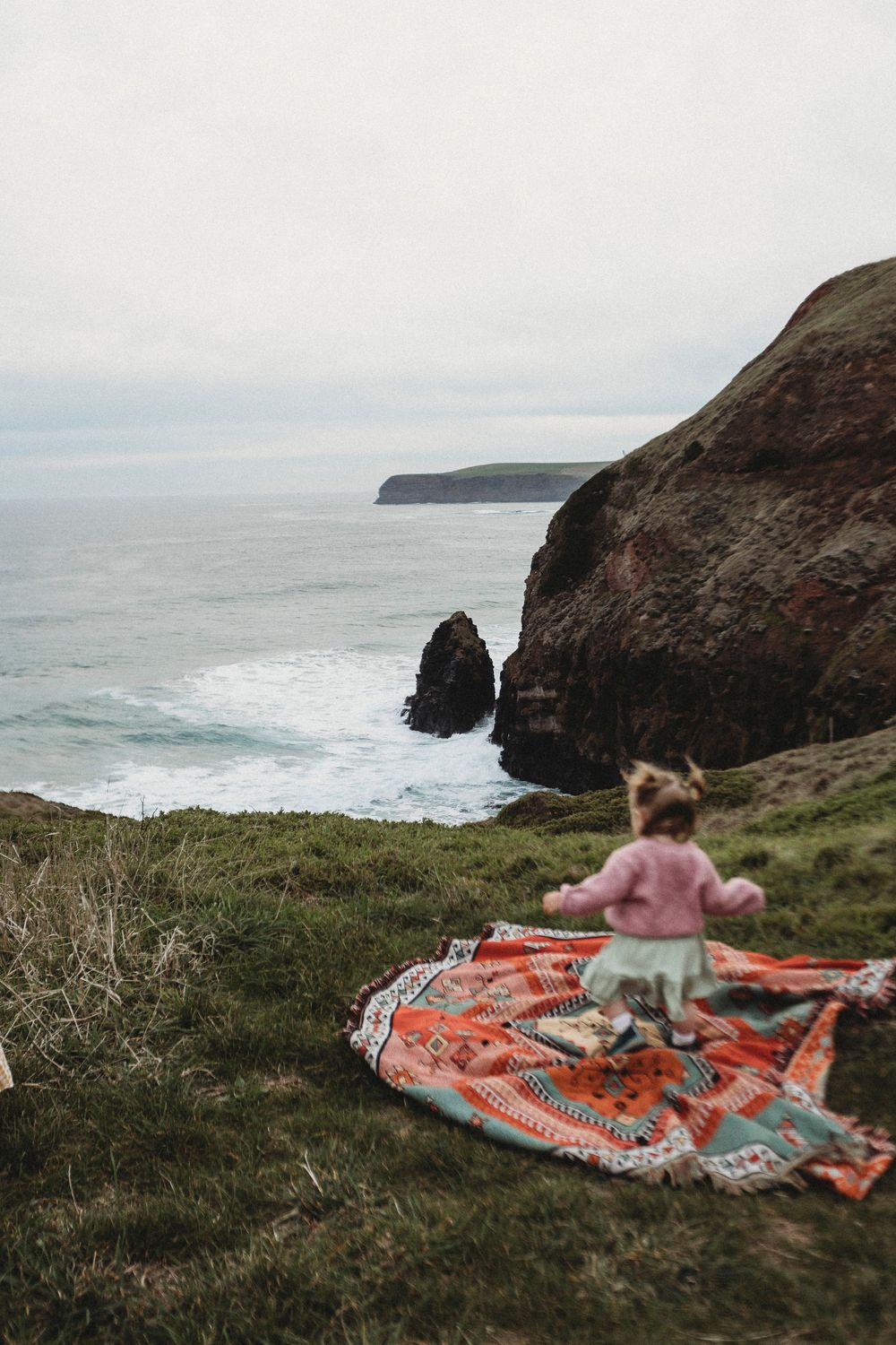 A young child stands on a colorful blanket on a coastal cliff, overlooking rocky sea stacks and ocean waves.
