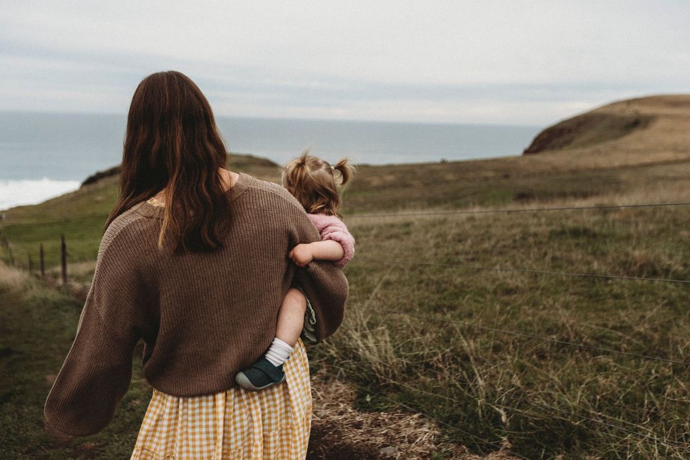 A mother holds her young child while gazing at a scenic coastal landscape with green hills and ocean views.