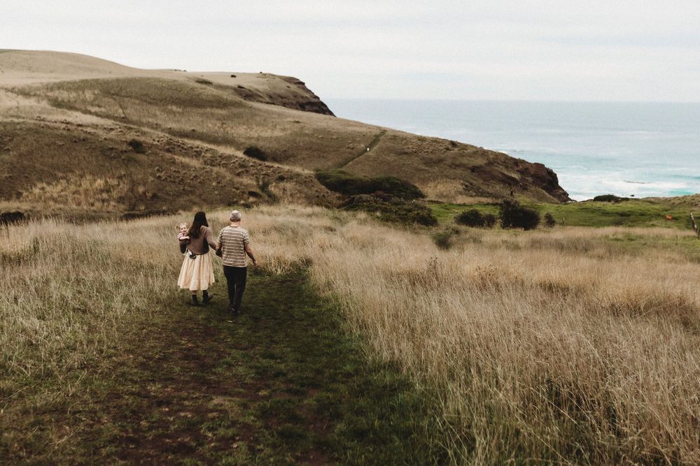 Couple walking hand in hand along a coastal trail through golden grass fields with ocean views.