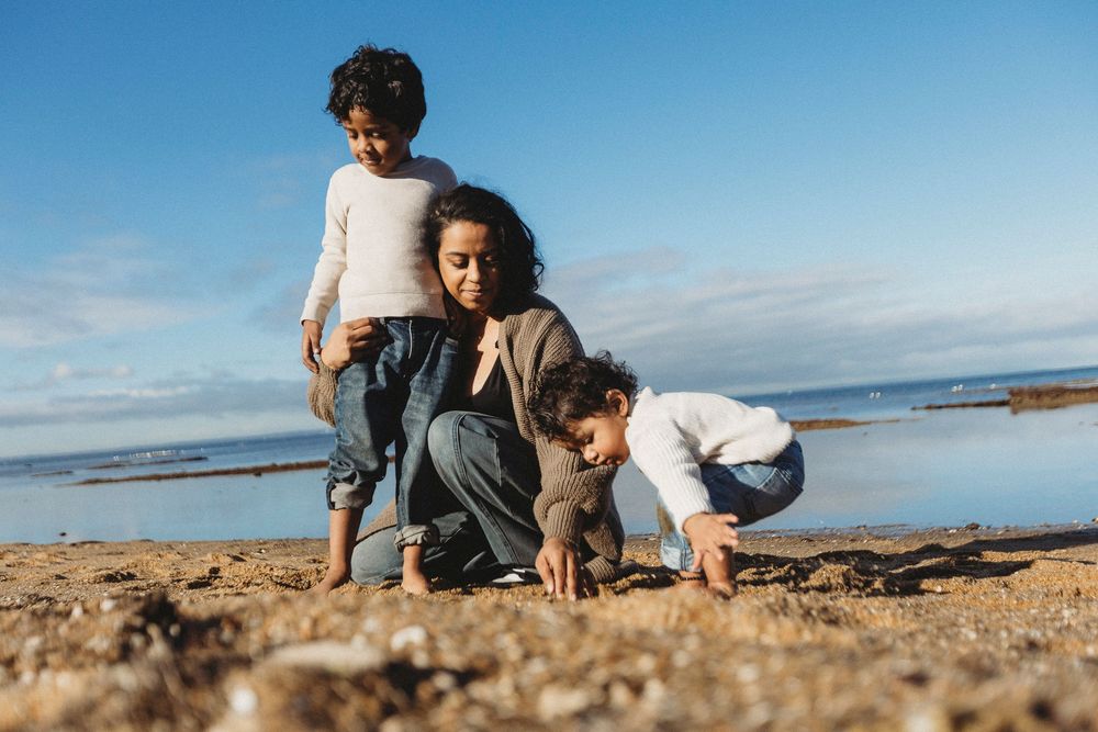 A mother and two young children playing on a sandy beach shore under a bright blue sky.