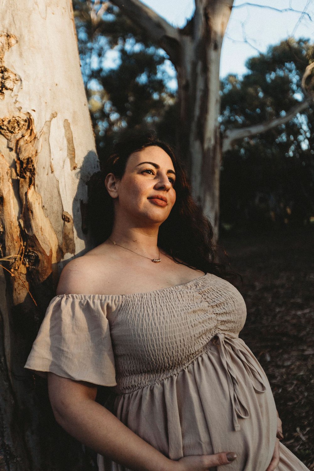 Pregnant woman in off-shoulder dress leaning against tree in golden hour light outdoors.