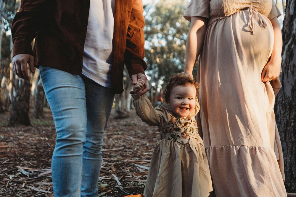 Happy toddler girl holds hands with parents during an outdoor maternity photoshoot among autumn trees.