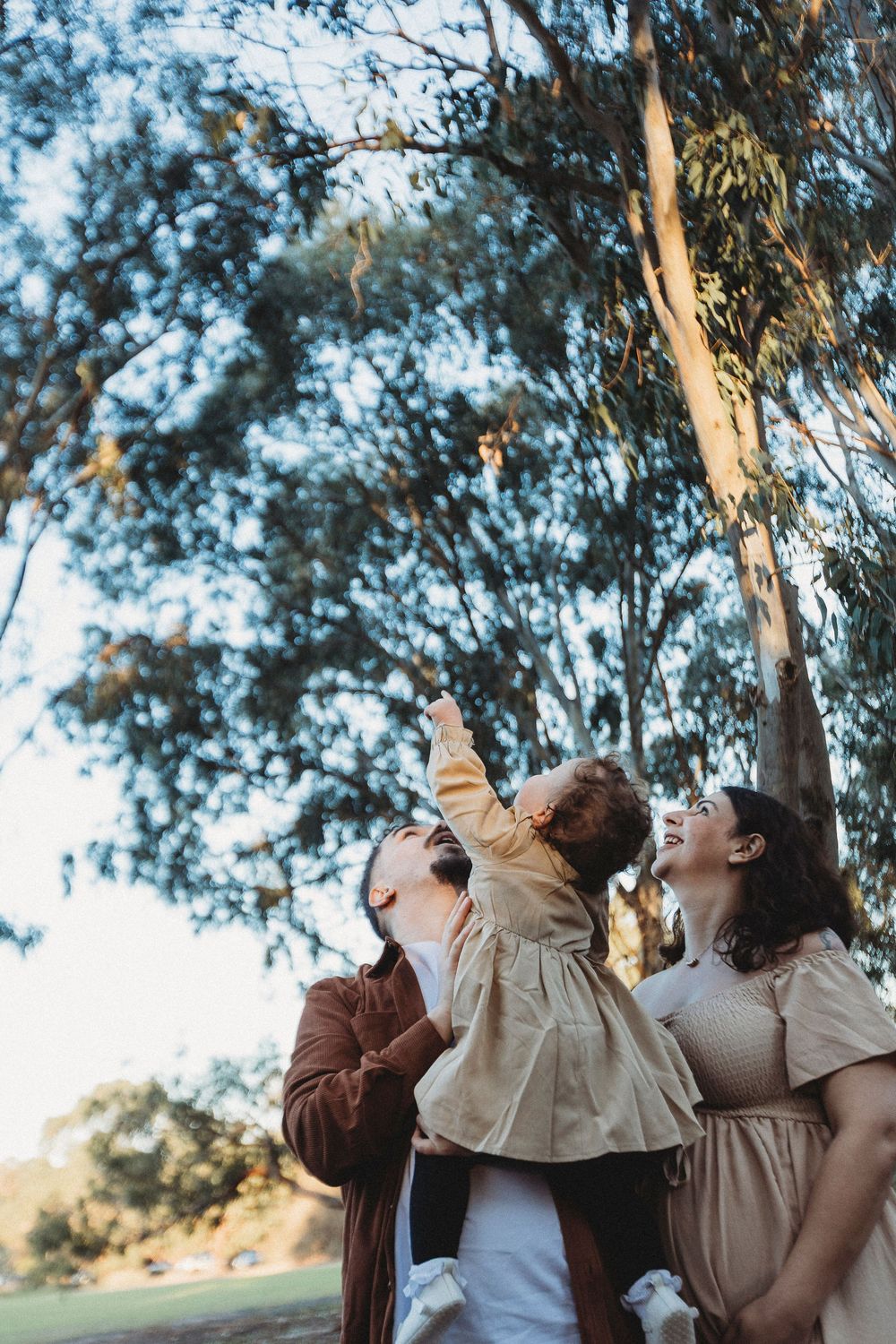 Family with toddler laughing outdoors under eucalyptus trees, child pointing up joyfully.
