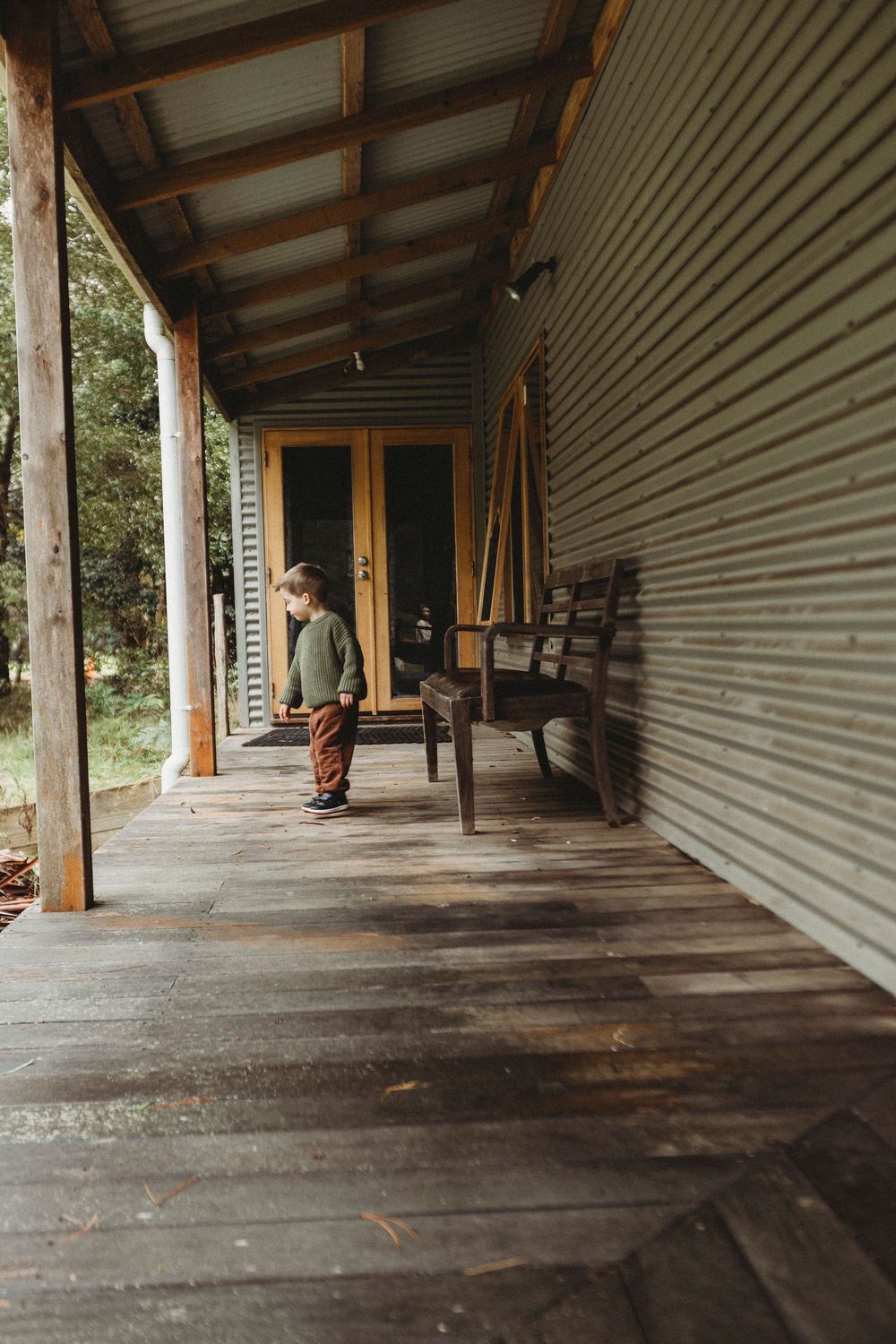 Young toddler standing on a rustic wooden porch of a corrugated iron cabin, with a wooden bench nearby.
