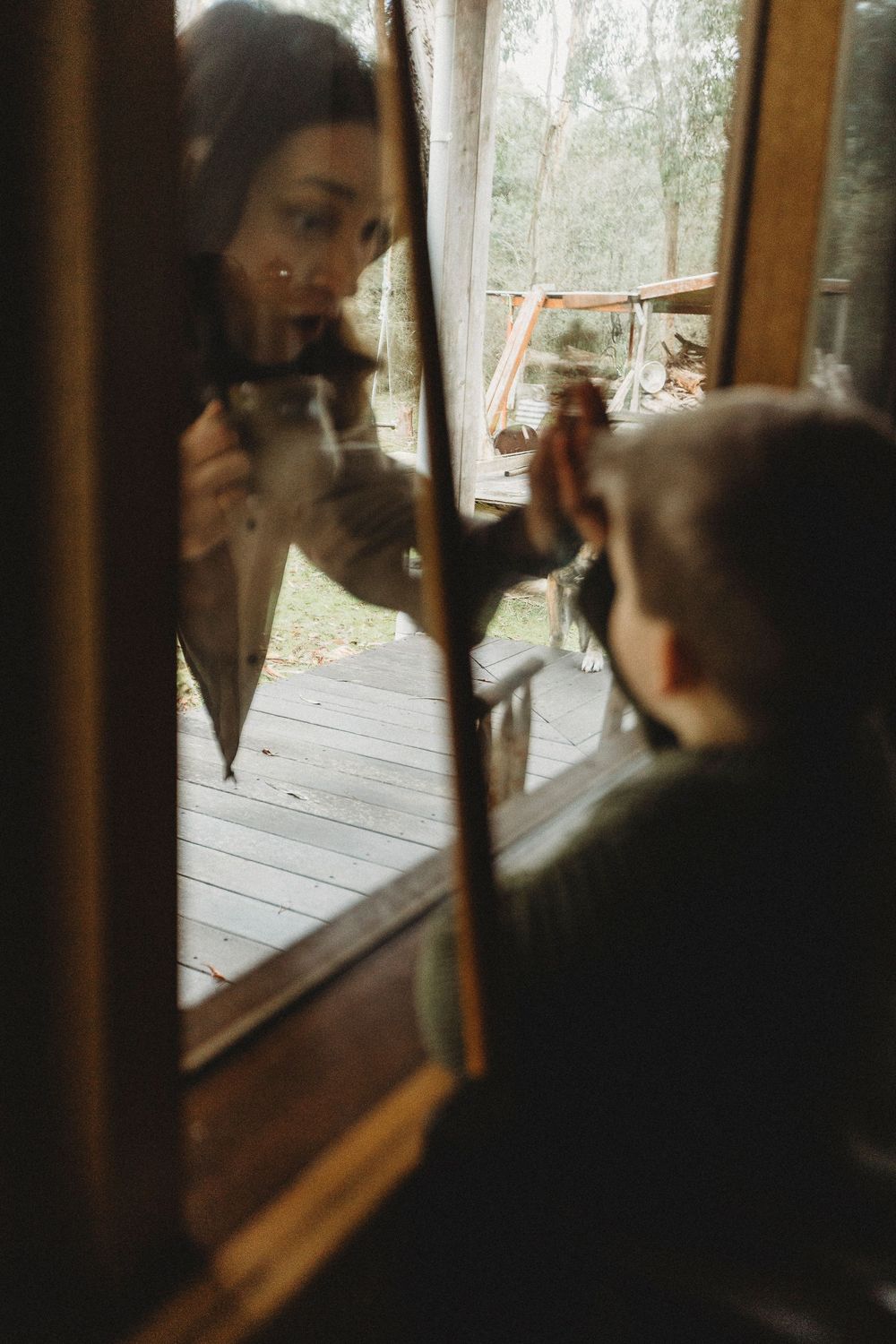 Woman and child interact through a rustic wooden window of a cabin surrounded by lush forest greenery.