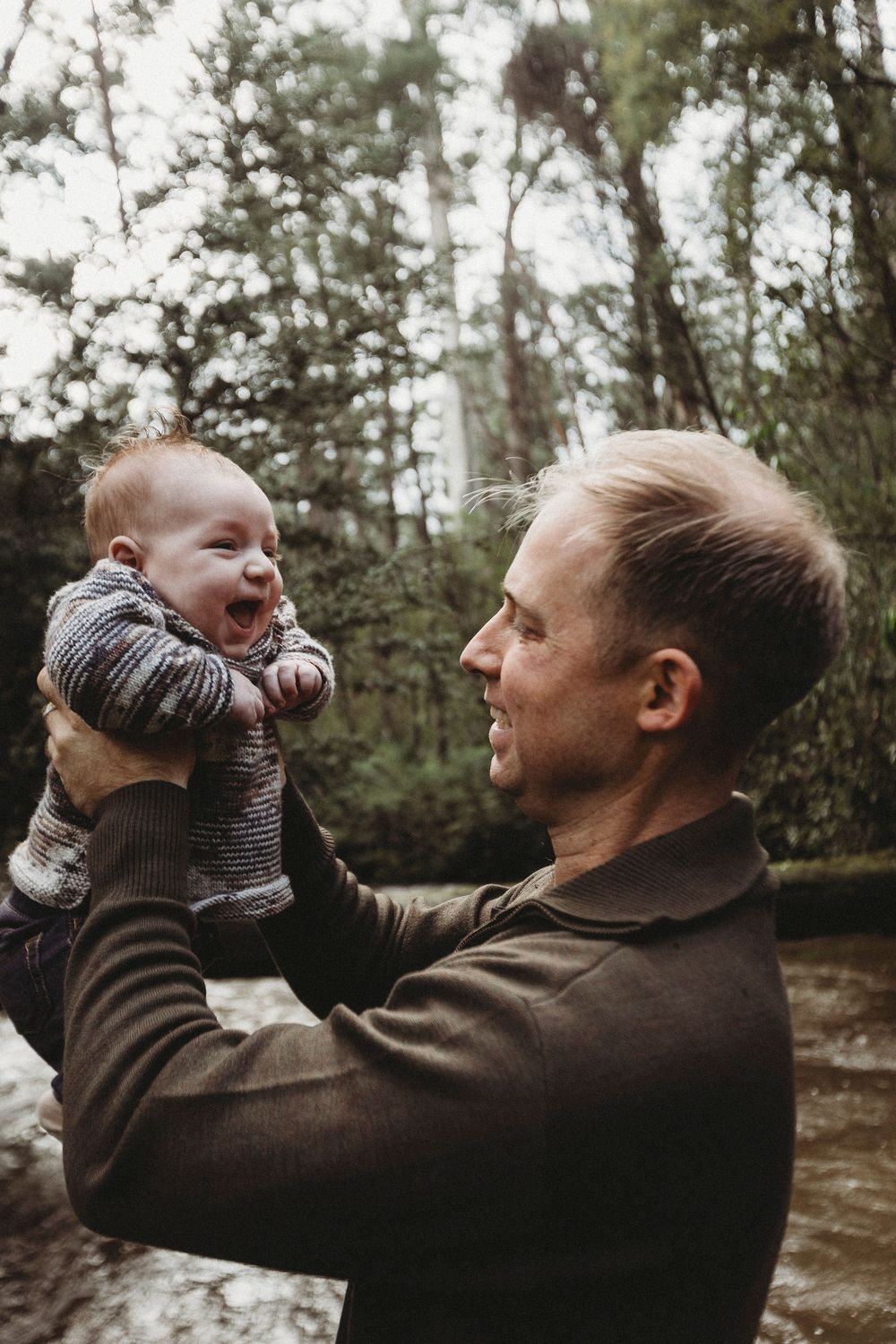 A father joyfully lifts his laughing baby in a lush forest setting near a stream.