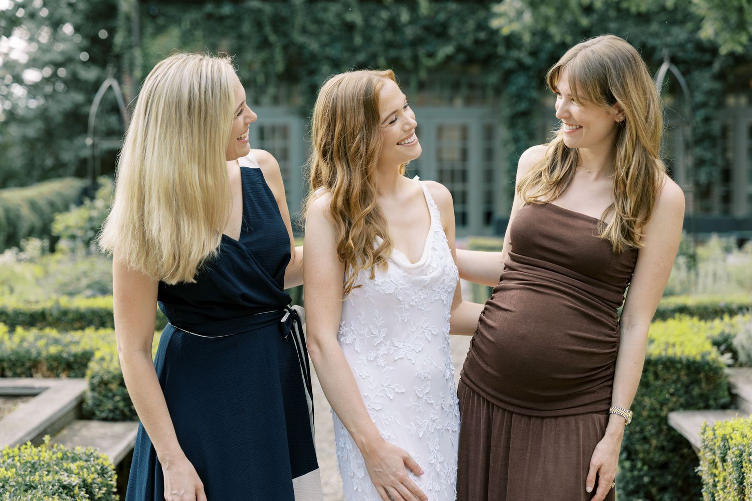 Three people stand together in a garden wearing coordinating formal dresses in navy, white and brown tones at Ninety Acres.