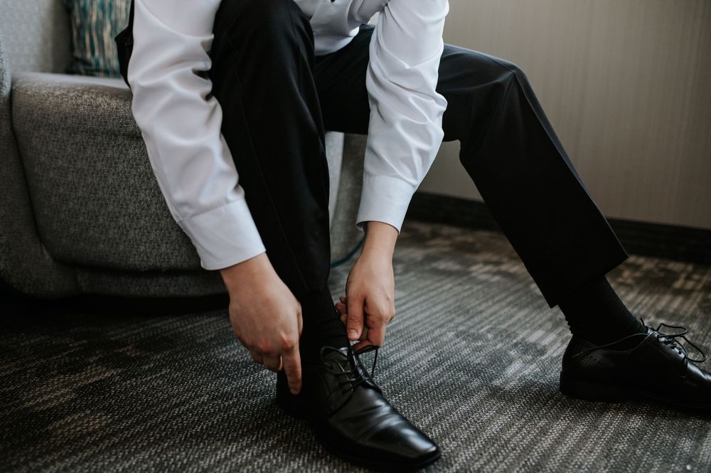 Someone in formal attire ties leather dress shoes while seated on a gray couch.