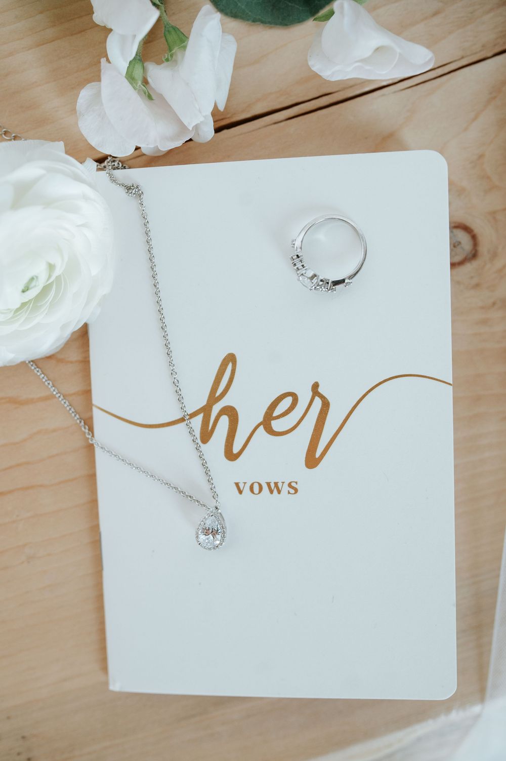 White wedding vow book with 'her vows' in gold text, ring and necklace on wooden surface.