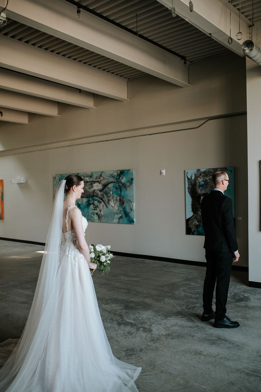 A wedding couple in formal attire stands in a modern industrial space with turquoise artwork on the walls.