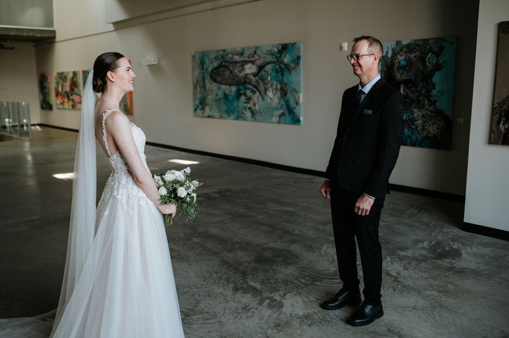 A couple in wedding attire stand in an art gallery with blue paintings on the wall.