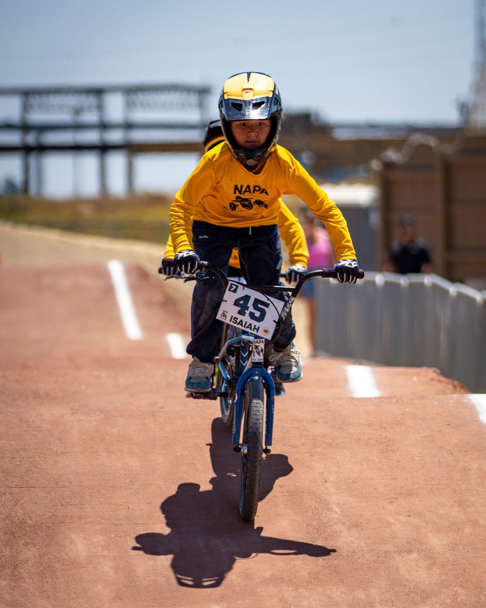 BMX rider performs jump sequence on dirt track wearing bright yellow jersey.