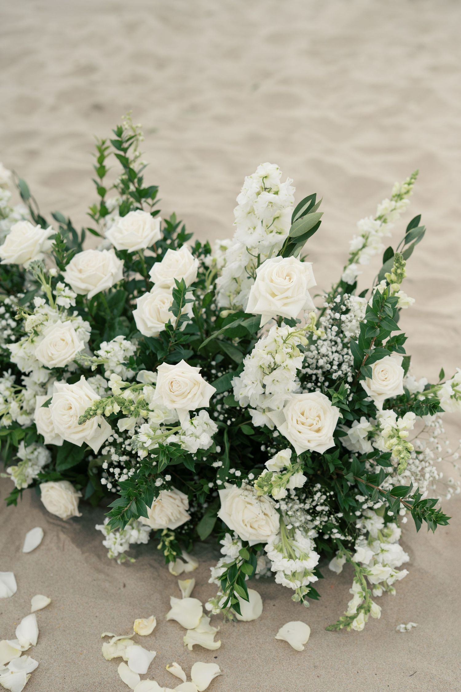 Elegant white roses and delicate baby's breath floral arrangement on sand with scattered petals.