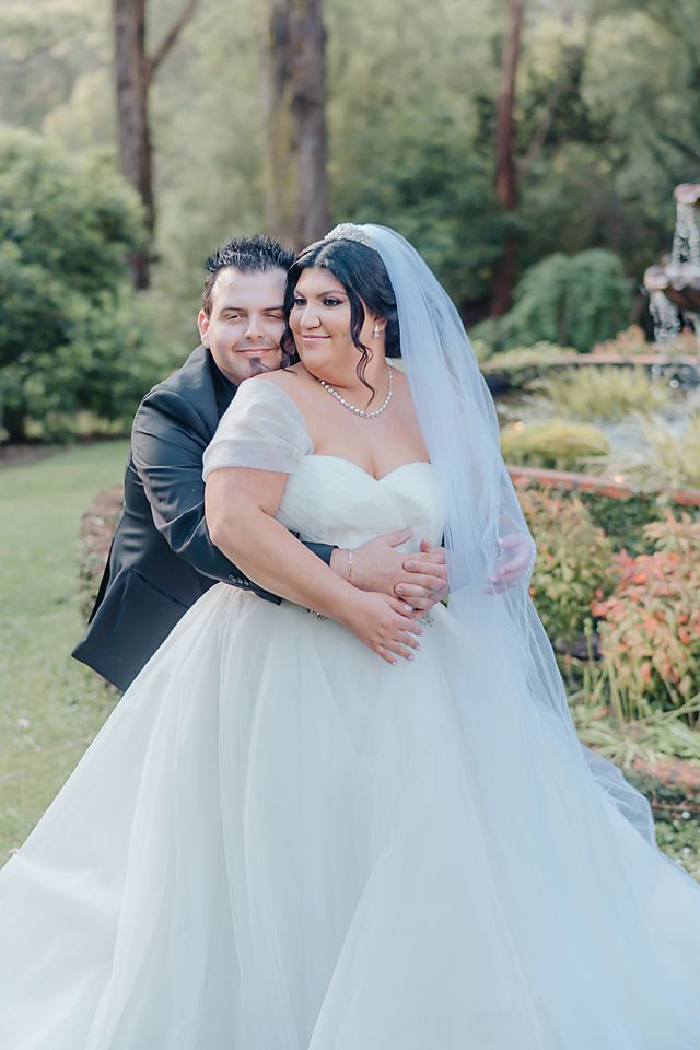A bride and groom embrace in an outdoor garden setting among tall trees and greenery on their wedding day.