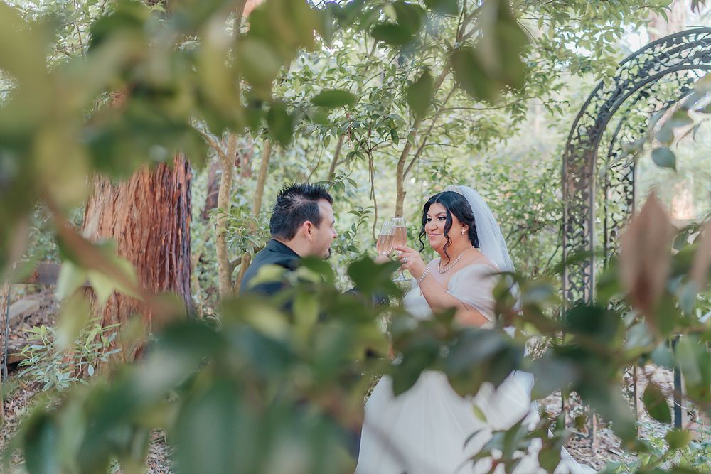 A couple in wedding attire shares a romantic moment in a lush garden setting with decorative arches.