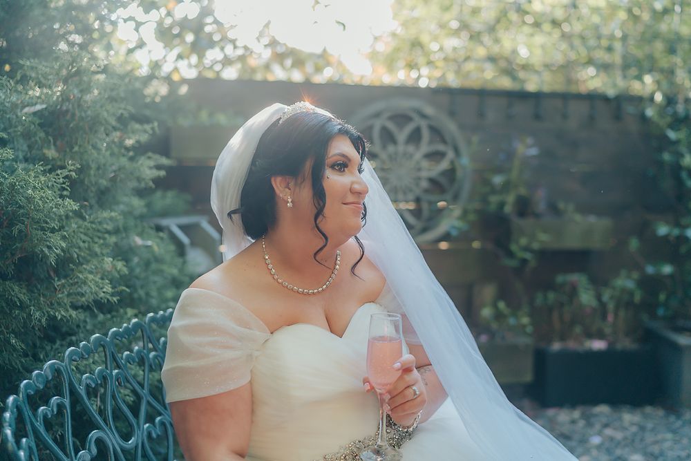 A bride wearing an off-shoulder wedding dress and long veil poses in a garden setting during golden hour.