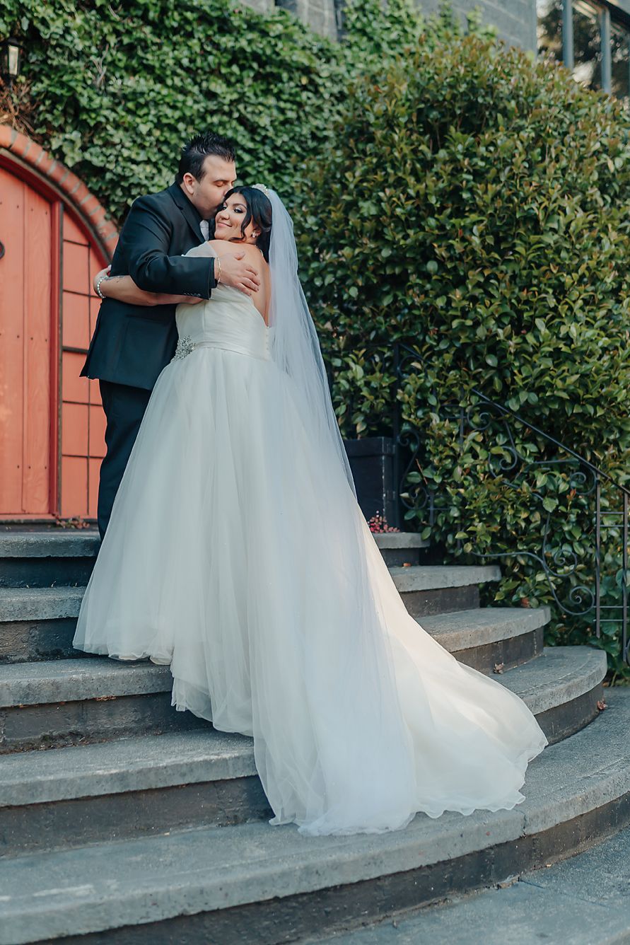 A romantic wedding couple in formal attire embracing on curved stone steps with ivy-covered walls behind them.