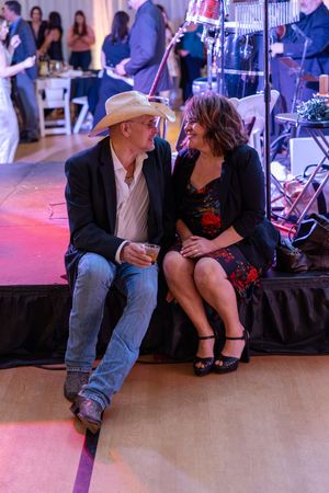 A couple in western attire sitting close together and laughing on the edge of a stage at a social event.