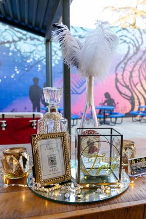 Elegant party decor display with white feather, crystal decanter, vintage frames and decorative glassware on mirrored tray.