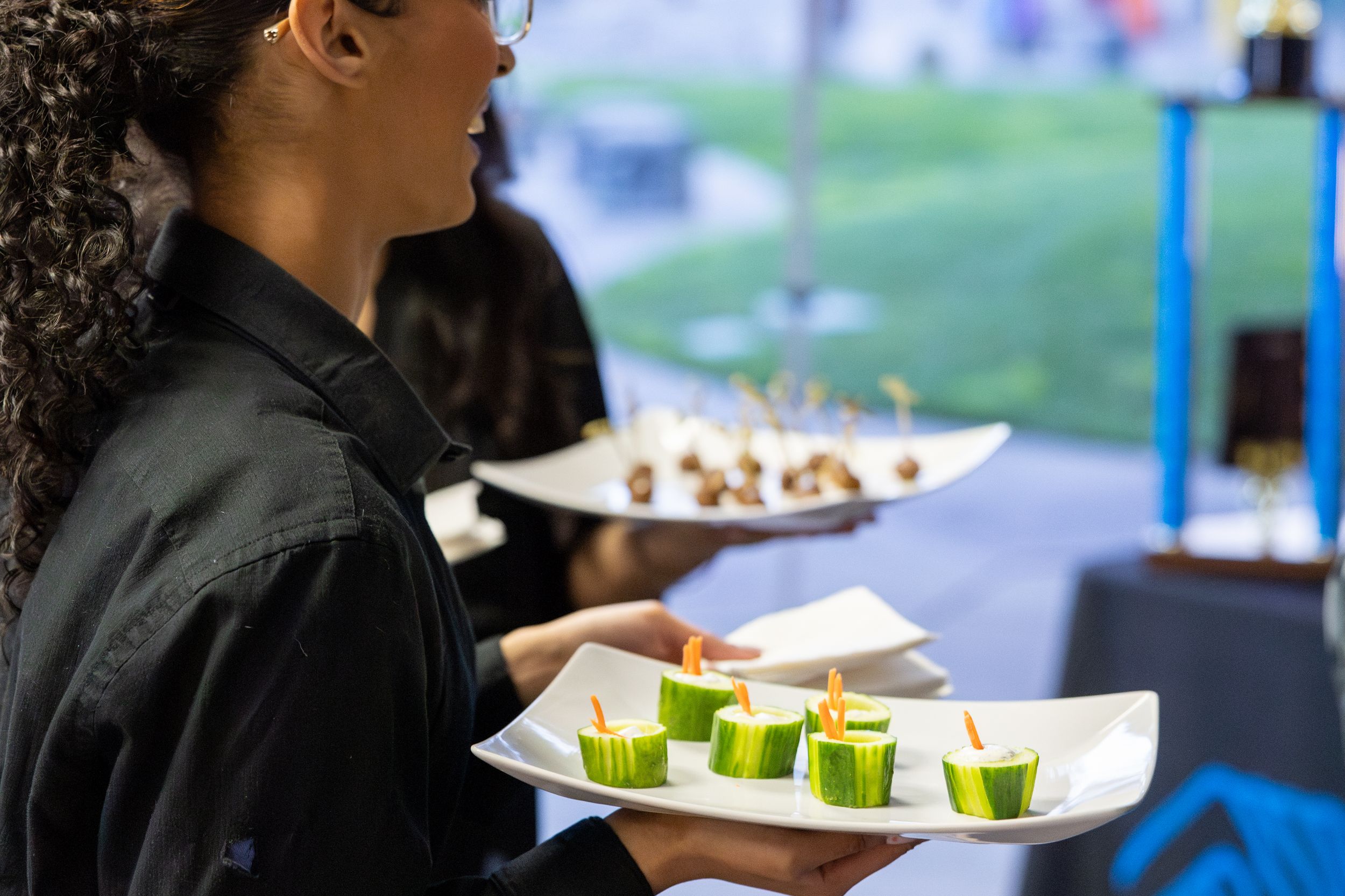 A close-up view of catering staff walking and serving food on white plates at an indoor event.