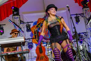 Performer in black corset, striped stockings, and orange jacket energetically sings on stage during outdoor festival performance.