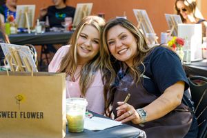 Two people smile while seated at an outdoor cafe table with drinks and a Sweet Flower shopping bag.