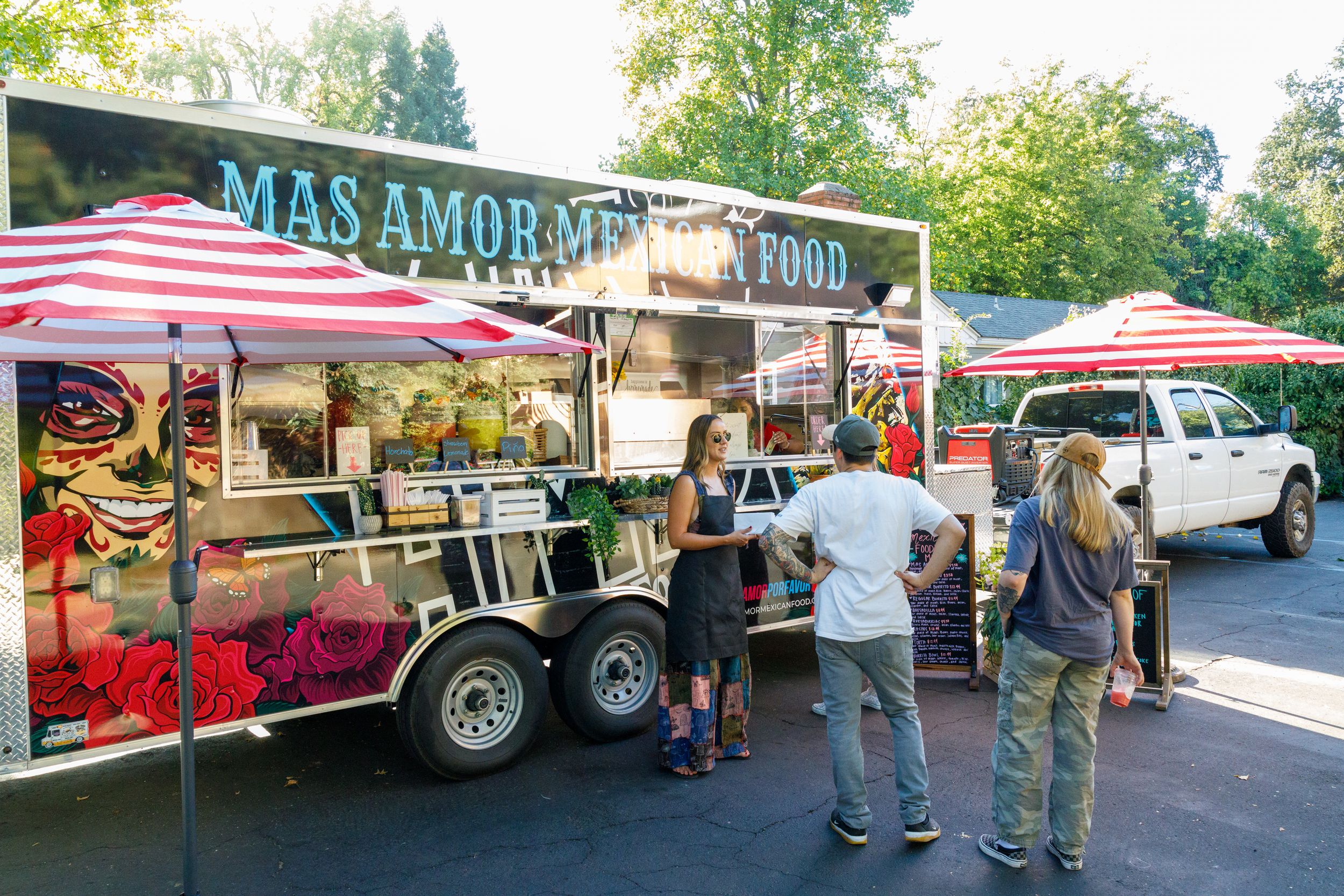 Customers line up at a colorful Mas Amore food truck with red striped umbrellas in a parking lot on a sunny day.