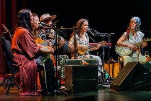Musicians perform together on stage with various instruments including strings and percussion during a live concert performance.