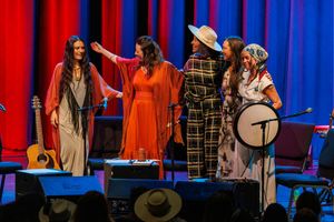 Performers in red and white robes stand on stage with musical instruments against a vibrant red and blue backdrop.