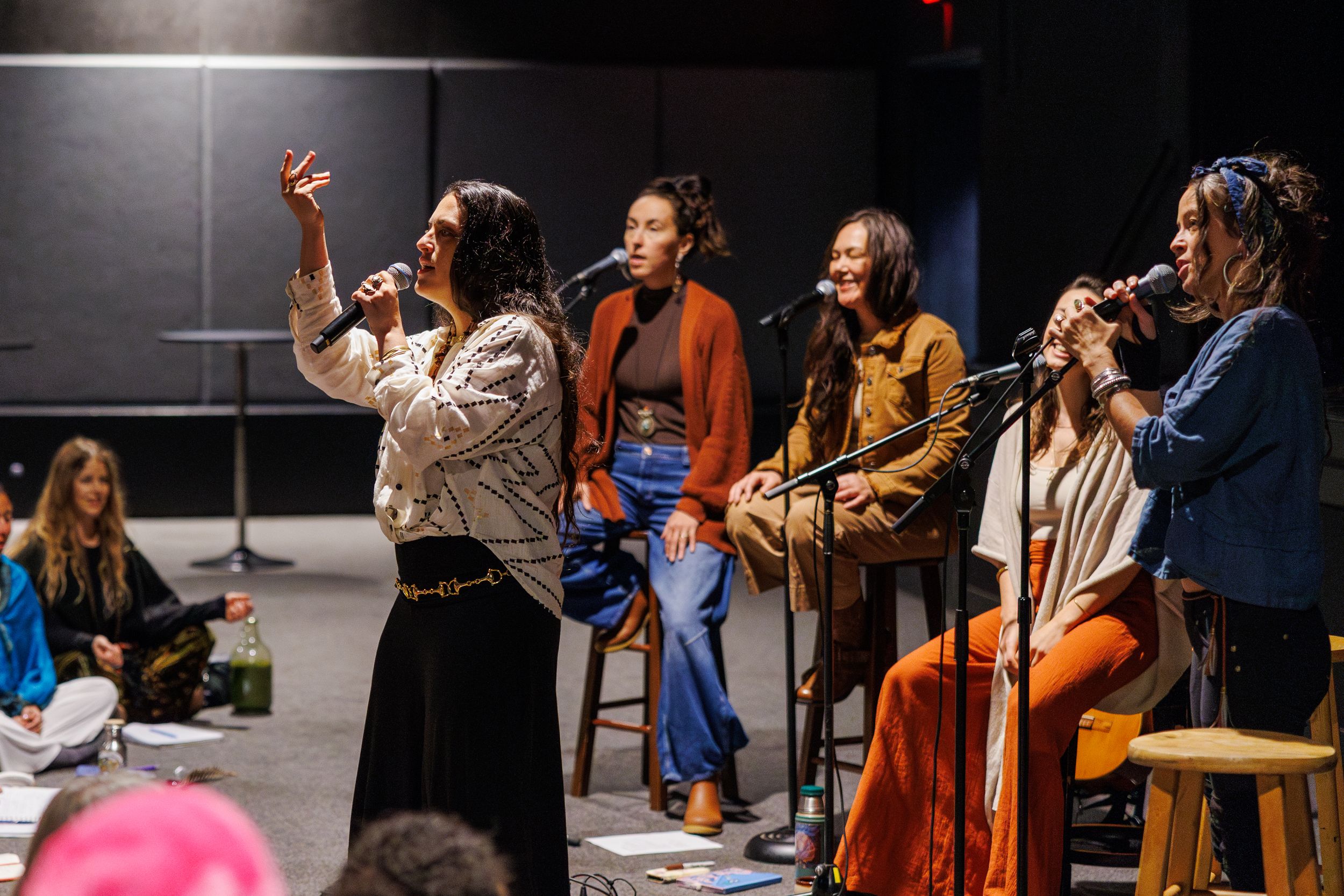 A group of musicians rehearse together in a dark studio space, some standing while others sit on wooden stools.