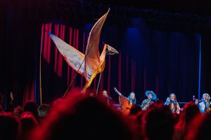 Illuminated origami bird prop glows on dark stage above concert crowd silhouettes with blue and red lighting effects.