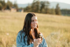 Someone wearing denim jacket holds wildflowers in golden field at sunset with mountains in background.