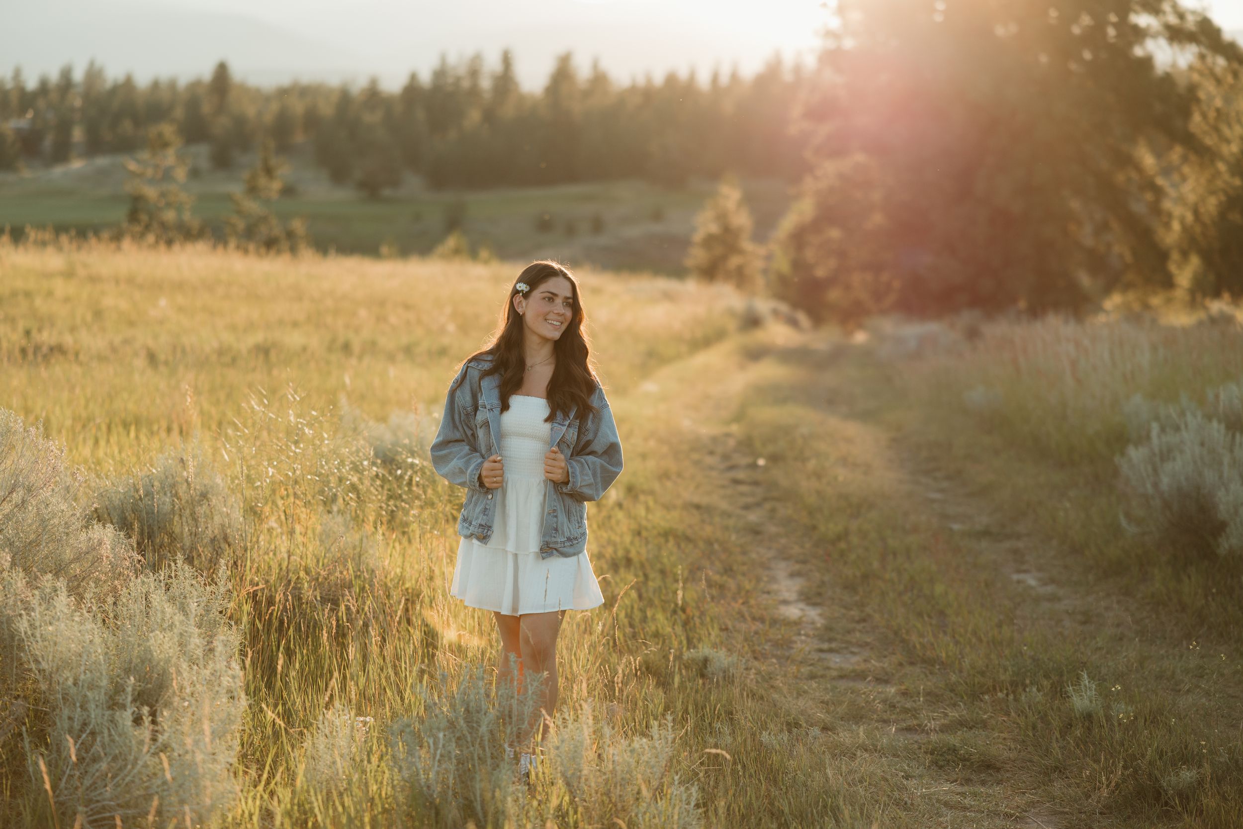 A woman standing in a golden grassy field at sunset gazing into the distance in a dreamy setting.