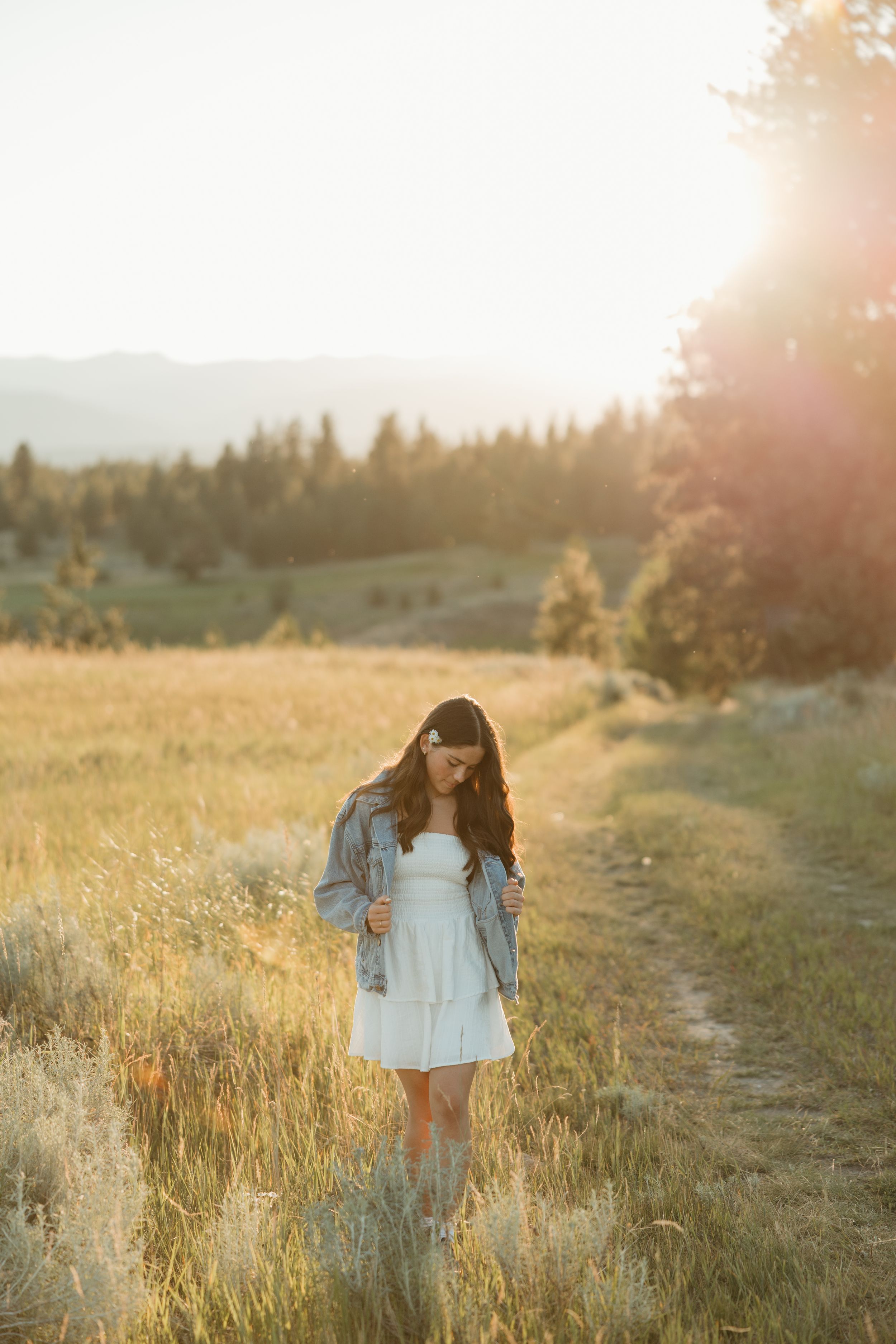 A person in a white dress and denim jacket stands in a golden meadow at sunset with mountains and forest in the distance.