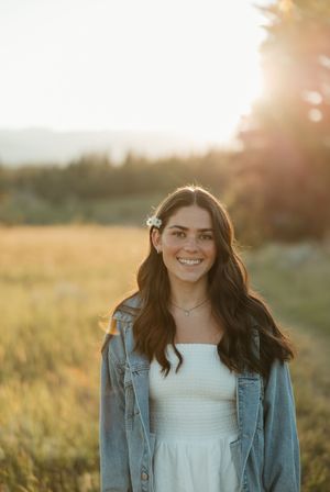 A person in a denim jacket and white top stands in a sunlit field during golden hour with sun flare behind them.