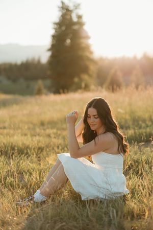 Woman in flowing white dress sits peacefully in golden sunlit meadow during sunset with mountains in background.
