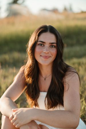 Portrait of a smiling person with long dark hair in white top sitting outdoors during golden hour.