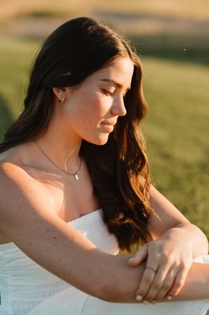 Woman in white dress sitting outdoors at sunset with golden light illuminating her long dark hair and delicate jewelry.