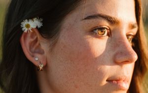 A white daisy adorns dark hair while golden sunlight illuminates freckled skin and an eye in profile view.
