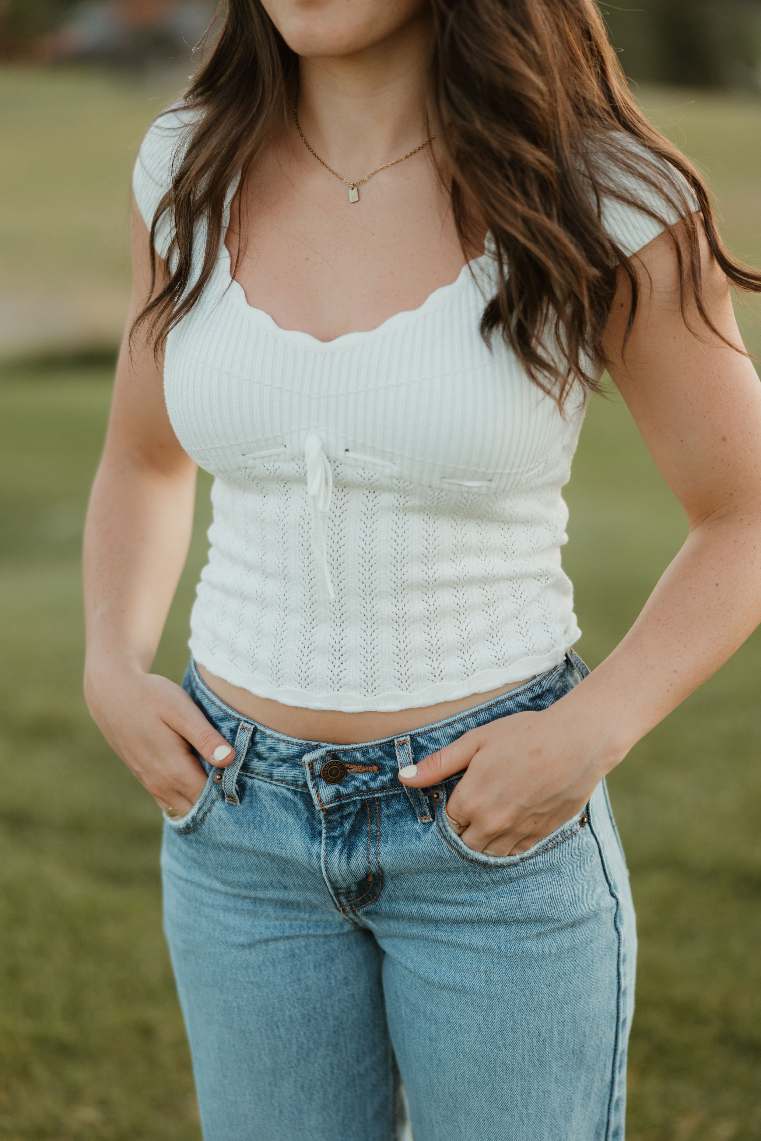 Woman wearing white ribbed crop top with scalloped neckline and light blue denim jeans in outdoor setting.