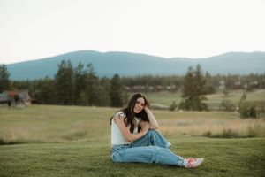 A casually-dressed person sits in a relaxed pose on green grass with mountains and trees in the background.