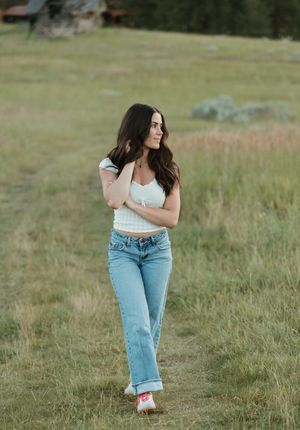 The model poses casually in a white top and light-wash jeans while walking through a grassy meadow.