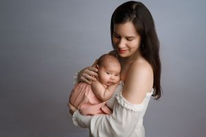 Tender portrait of mother and newborn baby embracing against neutral gray background in soft natural light.