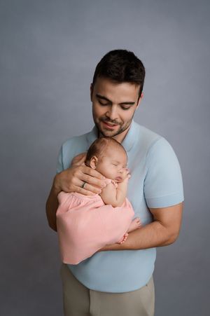 A tender moment captured as someone in a light blue shirt cradles a sleeping newborn baby wrapped in pink against a gray backdrop.