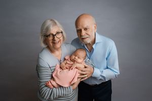Portrait of happy grandparents holding a sleeping baby in pink outfit against grey backdrop.