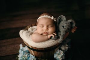 Sweet newborn rests peacefully in a wooden bowl with knit lovey toy and lace headband on dark wooden backdrop.