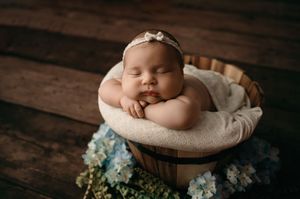 Sleeping newborn in wooden bowl surrounded by soft white fabric and blue hydrangea flowers on wooden floor.