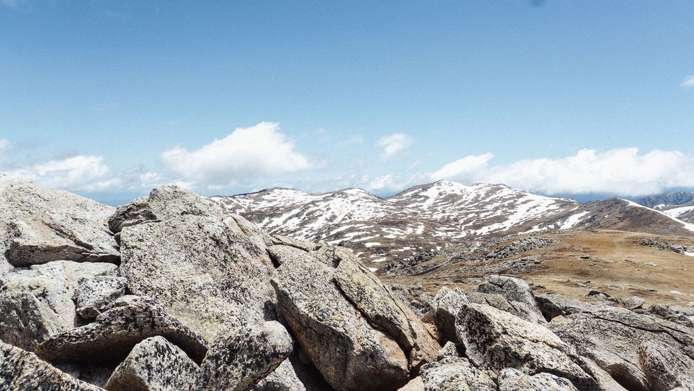 Rocky mountain summit with snow-capped peaks and boulders against a blue sky with white clouds in the distance.
