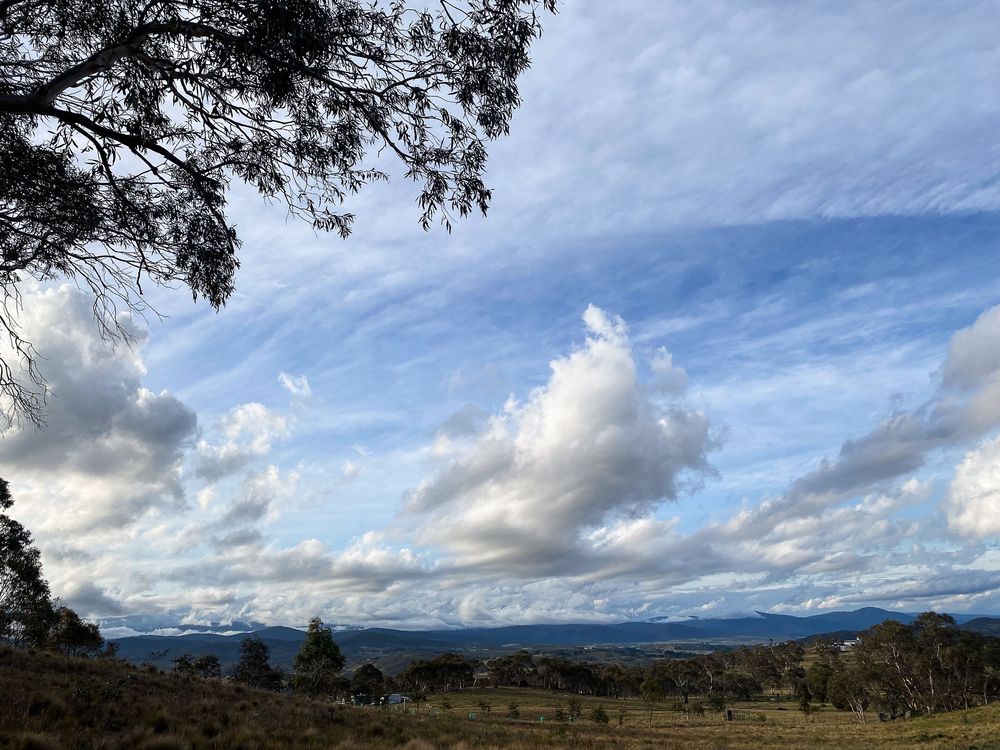 Dramatic panoramic landscape of rolling hills and mountains under a cloudy blue sky with scattered sunlight.