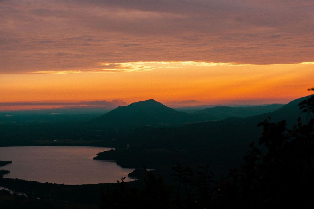 Stunning panoramic sunset view over mountainous coastline with orange and purple sky reflecting on calm ocean waters.